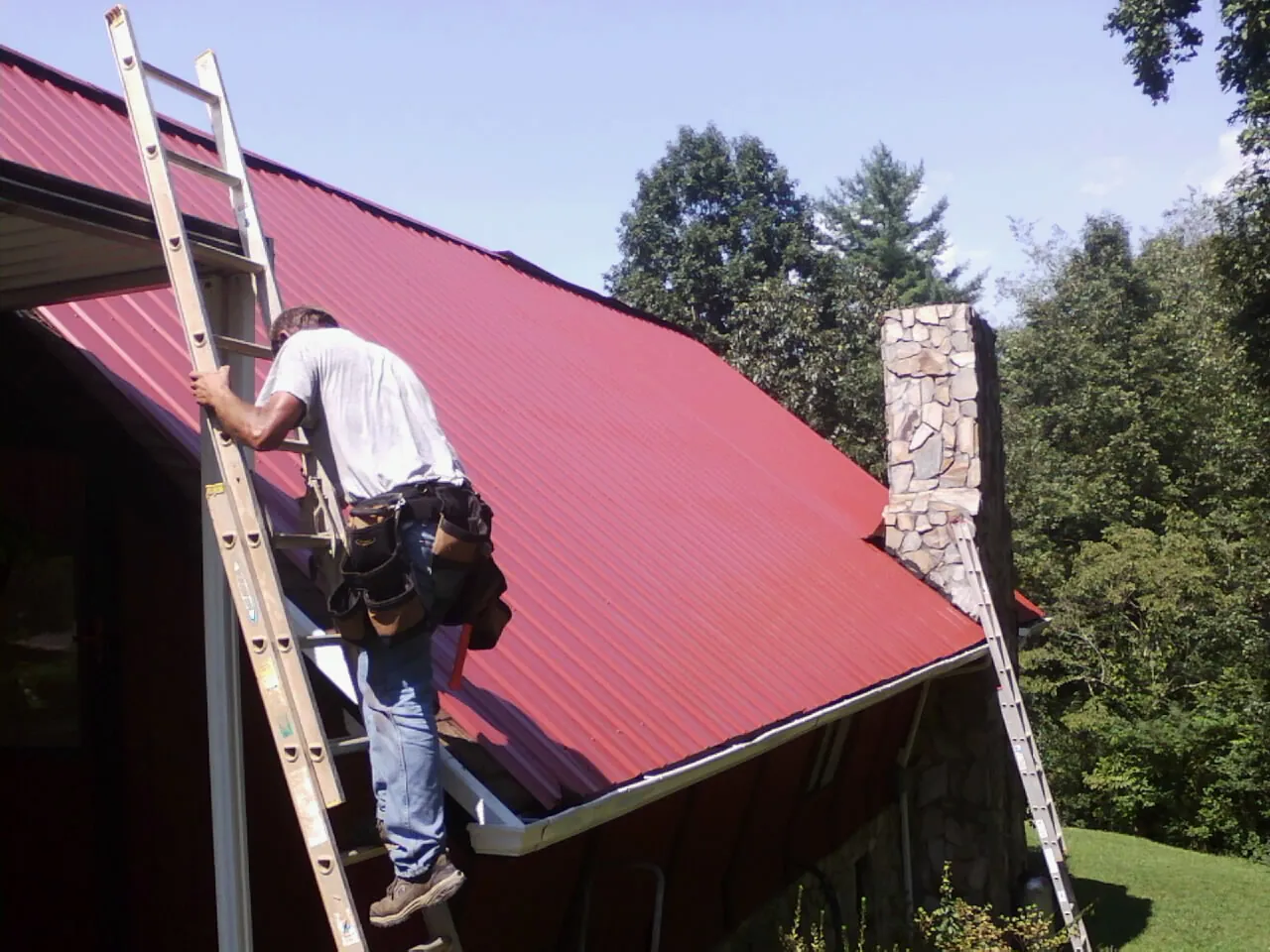 Roofer on a ladder beside a red metal roof and stone chimney — Soffit & Fascia Repair by Roofing Brothers in Allerton